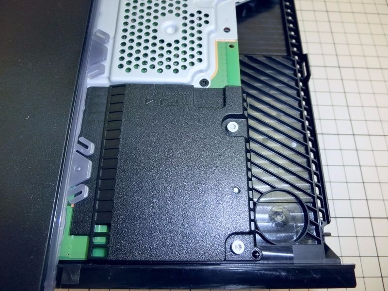 硬盘盖打开。这里我们发现了新版主机的第一个不同，原本一直覆盖到头的金属屏蔽板在CUH-1200主机中明显缩短，硬盘上方改用一块塑料盖板。这种设计虽然可以让硬盘部分的空间变得更紧凑，但对于硬盘的保护明显减弱。
值得一提的是，塑料盖板将旁边的12V接口进行了遮挡，避免了误触。