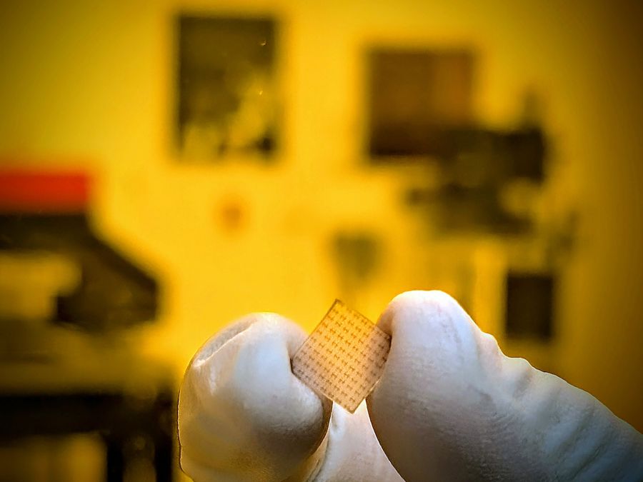 A small square with brown patterns is held between the finger and thumb of a white-gloved hand.